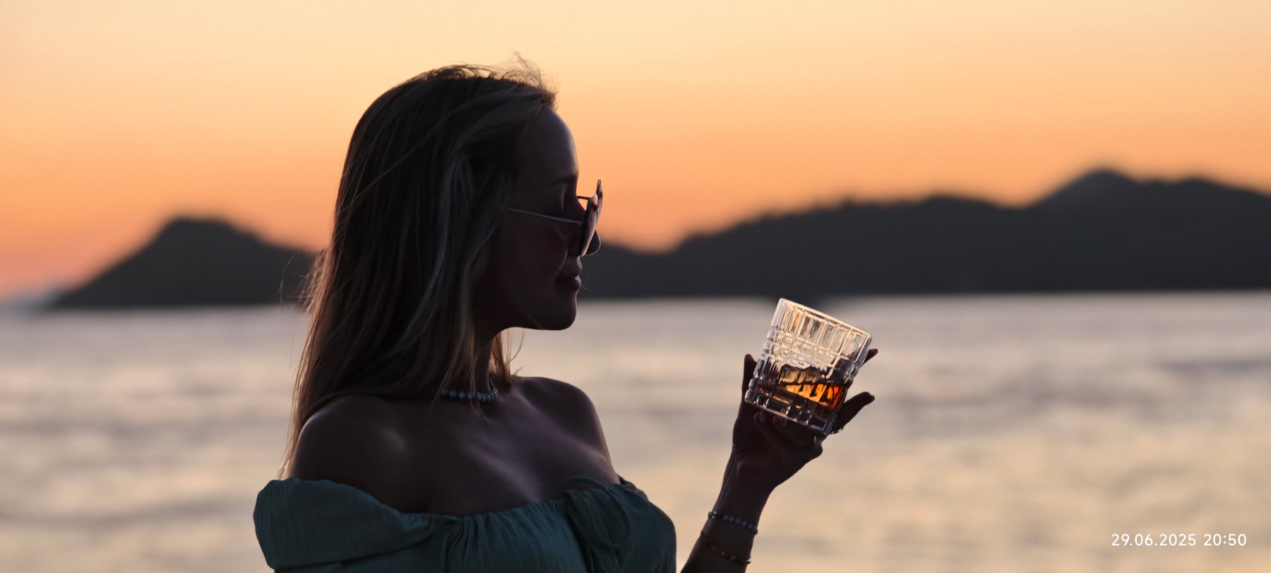 Elegante Frau genießt Whiskey aus einem strukturierten Whiskeyglas bei Sonnenuntergang am Meer